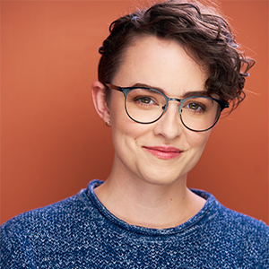 Erica Simpson headshot - Hanover Theatre and Conservatory - Hanover Theatre and Conservatory Portrait of a woman with short curly hair, glasses, and a blue knit sweater, against an orange backdrop (headshot).