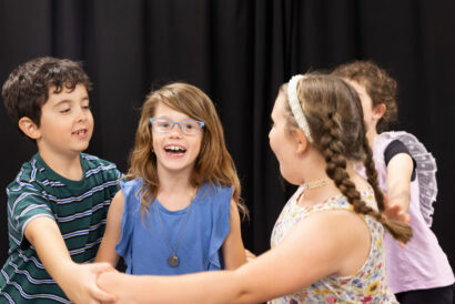 Folk Fairytale Grades 2-3 - Hanover Theatre and Conservatory Four children dance together on a stage with a dark curtain backdrop, holding hands and smiling.