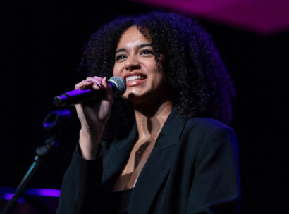 Female singer on stage, mid-performance, holding a microphone and smiling under purple stage lighting.