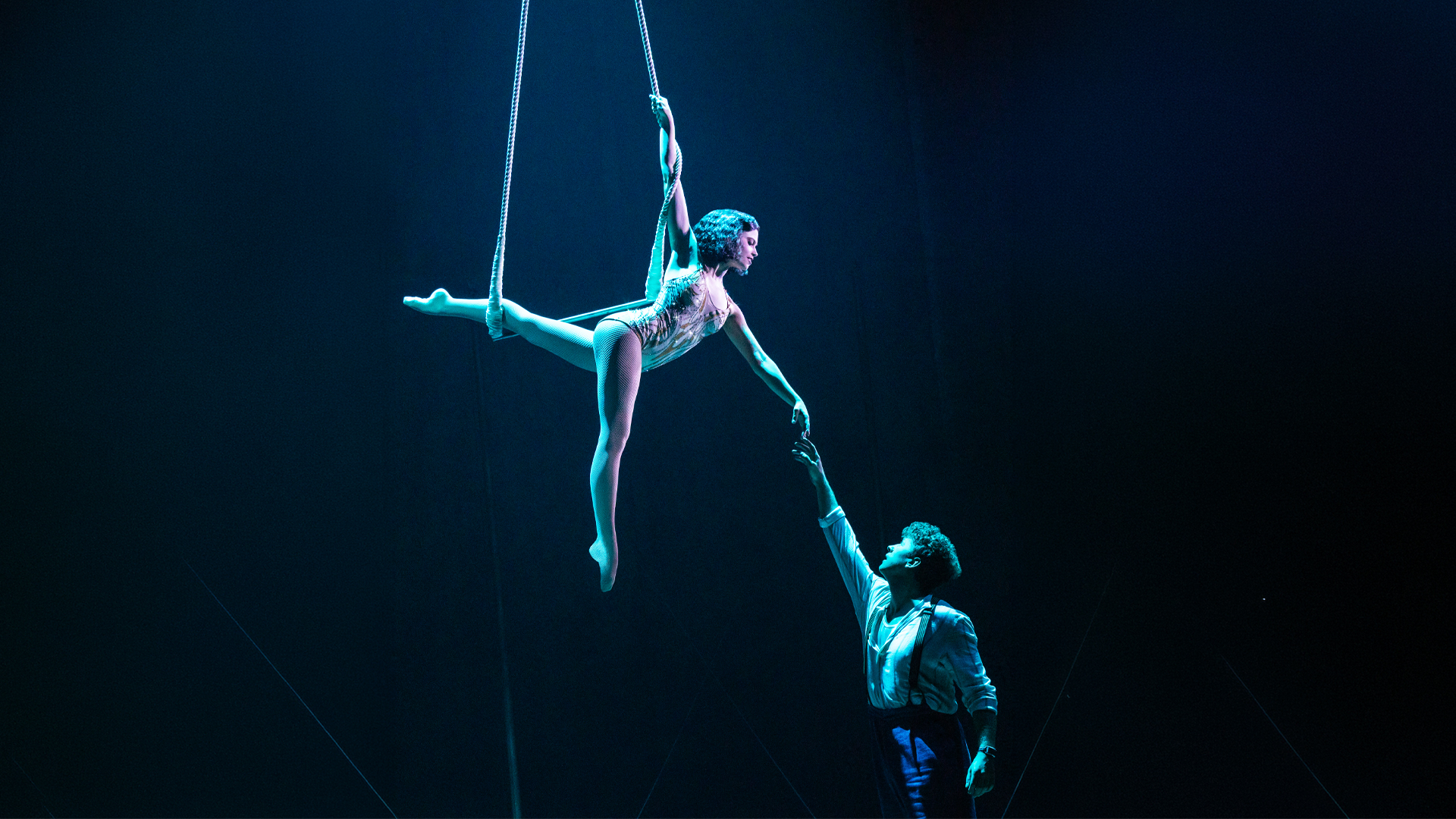 Aerialist in a leotard swings from ropes in a dark theater, with a male handler reaching up to help beneath her.