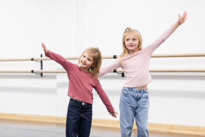Imaginative Play Grades K-1 - Hanover Theatre and Conservatory Two young girls stand at a ballet barre with arms stretched upward, smiling in a bright studio.