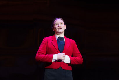 Intro to Singing Musical Theatre 2 Grades 4-7 - Hanover Theatre and Conservatory Female performer on stage wearing a red blazer and headset microphone, speaking or singing to an audience.