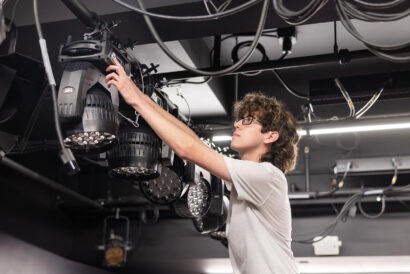 Intro to Technical Theatre Teens Adults - Hanover Theatre and Conservatory Person wearing glasses adjusts a large stage lighting fixture among cables overhead in a dim studio space