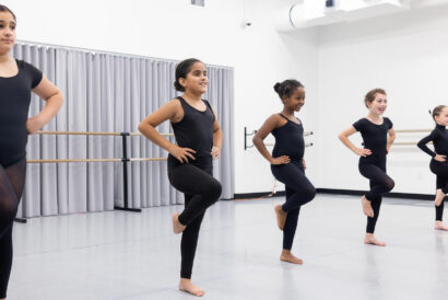 Jazz 2 Ages 10+ - Hanover Theatre and Conservatory Group of young girls in black leotards balancing on one leg with hands on hips in a ballet studio by a barre.