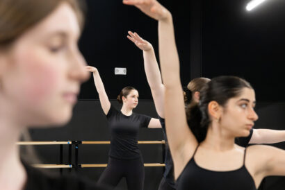Jazz 3 Ages 10+ - Hanover Theatre and Conservatory Dancers in black leotards practice ballet at a barre in a studio, arms raised in a synchronized pose, blurred foreground dancer on left.