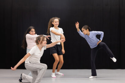 Lets Make A Play Grades 2-5 - Hanover Theatre and Conservatory Group of young dancers mid-motion in a studio with a black backdrop, posing dynamic dance moves and smiles.