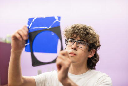 Lighting 1 Teens Adults - Hanover Theatre and Conservatory Young man with curly hair and glasses holds up a blue plastic panel in a cluttered room with a lavender wall.