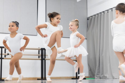 Pre-School Dance Ages 3-4 - Hanover Theatre and Conservatory Young ballet students in white leotards and tutus jump with hands on hips in a studio with bars along the wall.