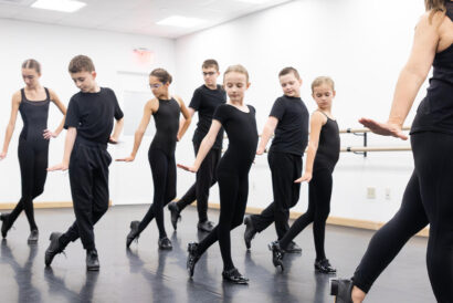 Primary Tap 2 Ages 10-12 - Hanover Theatre and Conservatory Group of kids in black leotards practicing ballet in a studio with an instructor at the right edge overseeing them