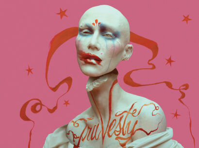 Avant-garde sculpture of a pale head and upper torso with blue eye makeup, red lips, pink background, orange ribbon swirls and stars.