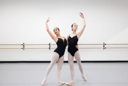 Senior Youth Ballet Company Invitation Only - Hanover Theatre and Conservatory Two female ballet dancers in black leotards and pink tights pose en pointe in a studio with a wooden barre behind them.