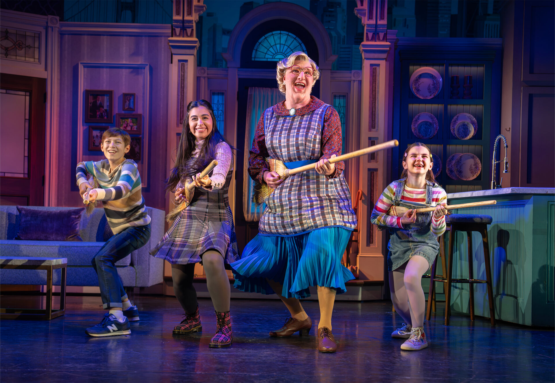 Group of four performers dancing on a theater stage with broom props in a bright kitchen set, smiling and energetic.