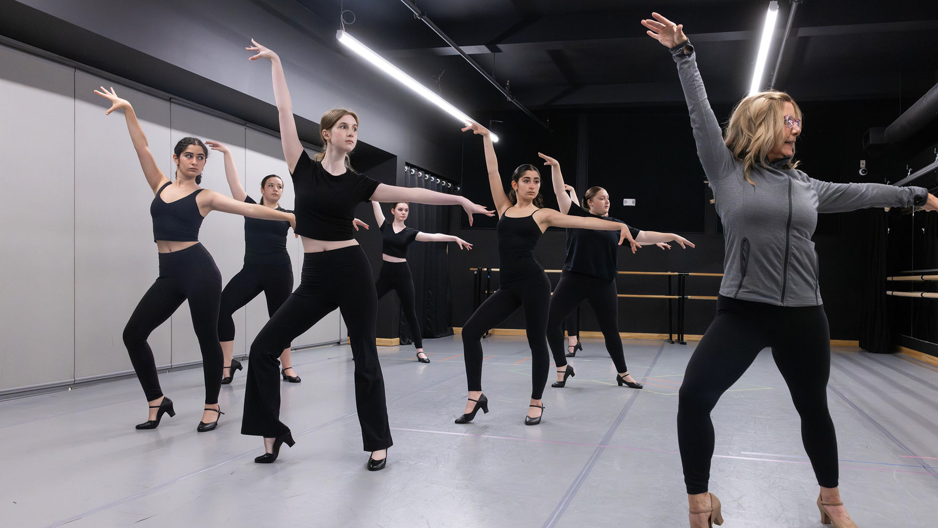 Group of dancers in a studio performing a synchronized pose with arms raised and one instructor at the front right guiding them.