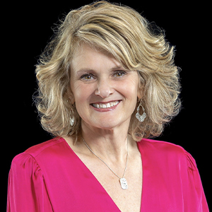 Sharon Hart headshot 2026 - Hanover Theatre and Conservatory - Hanover Theatre and Conservatory Portrait of a smiling middle-aged woman with curly blonde hair, wearing a pink blouse, silver earrings, and a pendant necklace against a black background.