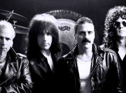 Four male rock musicians in leather jackets posing in a studio with a drum kit behind them, black-and-white photo.