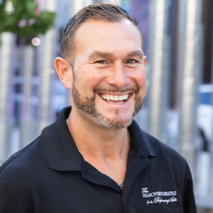 Dan G headshot - Hanover Theatre and Conservatory - Hanover Theatre and Conservatory Smiling man with short hair and beard wearing a black polo shirt, outdoors in an urban setting.