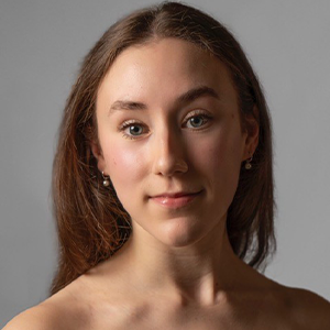 Grace Armour Headshot - Hanover Theatre and Conservatory - Hanover Theatre and Conservatory Close-up portrait of a young woman with straight brown hair looking at the camera against a gray background.