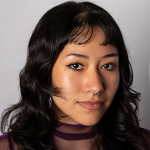 Sarah Malone Headshot - Hanover Theatre and Conservatory - Hanover Theatre and Conservatory Portrait of a young woman with dark wavy hair, wearing a purple sheer top and a septum piercing, looking at the camera.
