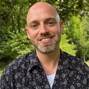 Wes Hunter headshot - Hanover Theatre and Conservatory - Hanover Theatre and Conservatory Smiling bald man with a trimmed gray beard outdoors, wearing a dark geometric-pattern shirt.