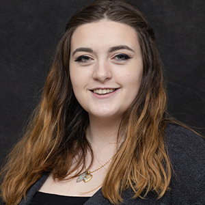 Allyson Kinch headshot - Hanover Theatre and Conservatory - Hanover Theatre and Conservatory Portrait of a smiling young woman with long brown hair, wearing a dark top and cardigan against a dark backdrop, with a subtle necklace.