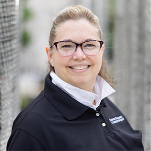 Maria S headshot - Hanover Theatre and Conservatory - Hanover Theatre and Conservatory Smiling woman with glasses, wearing a dark polo shirt, standing outdoors beside a chain-link fence.