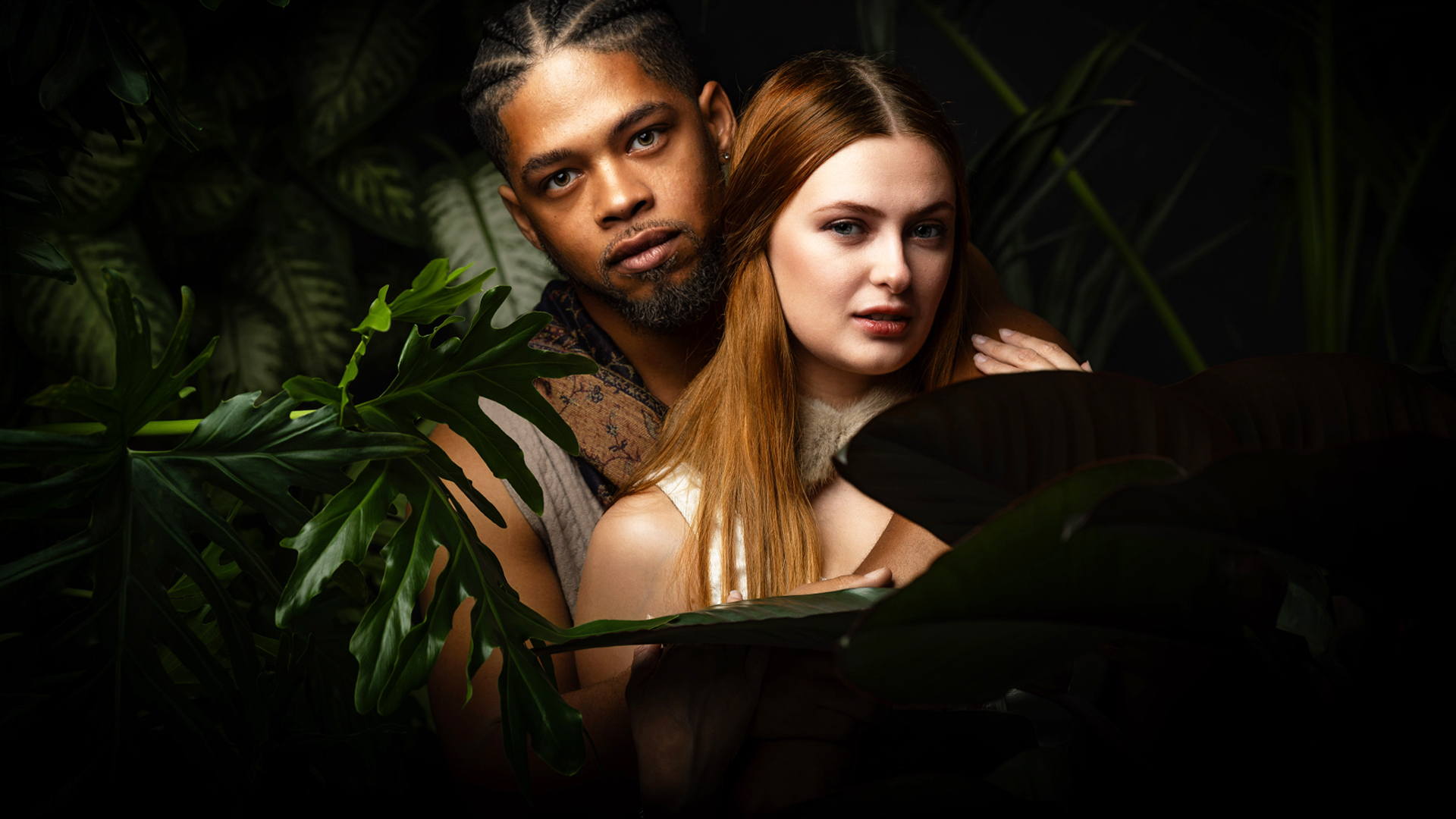 Couple nestled among tropical plants, the man behind the woman as they look at the camera.