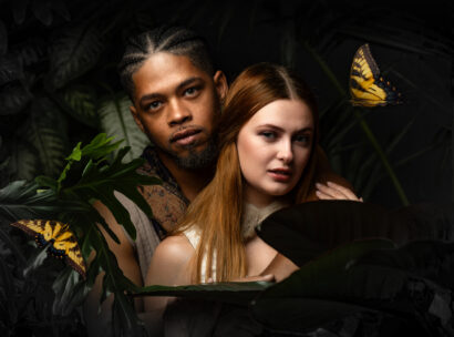 Couple in a tropical setting embraced among dark jungle leaves, with two yellow butterflies nearby on the edges of the frame.