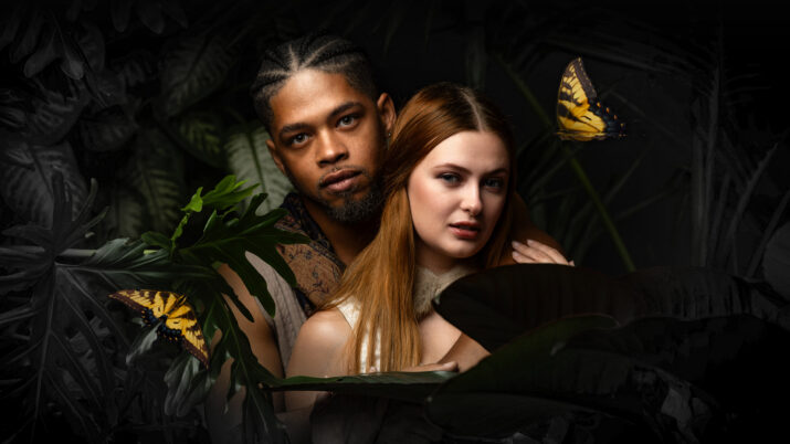 Couple in a tropical setting embraced among dark jungle leaves, with two yellow butterflies nearby on the edges of the frame.
