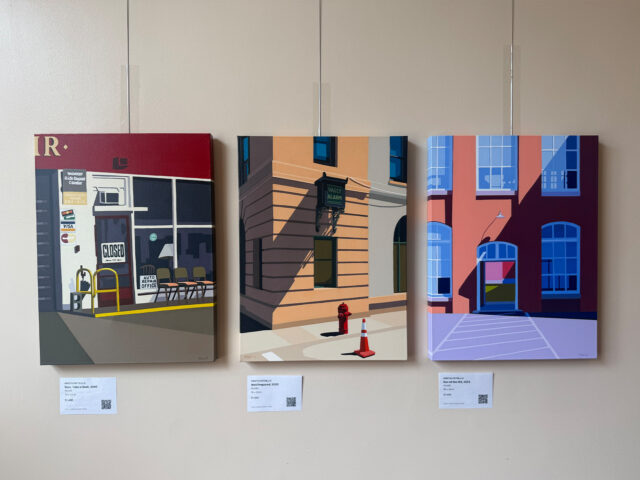 Triptych of three cityscape paintings mounted on a beige wall: left storefront with 'CLOSED' sign, middle brick building with a vault alarm, right modern pink building with blue windows.