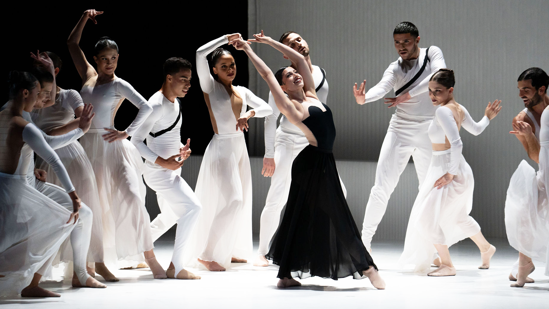 Group of dancers in white costumes with one dancer in a black dress performing a coordinated pose on stage, with graceful arm movements.